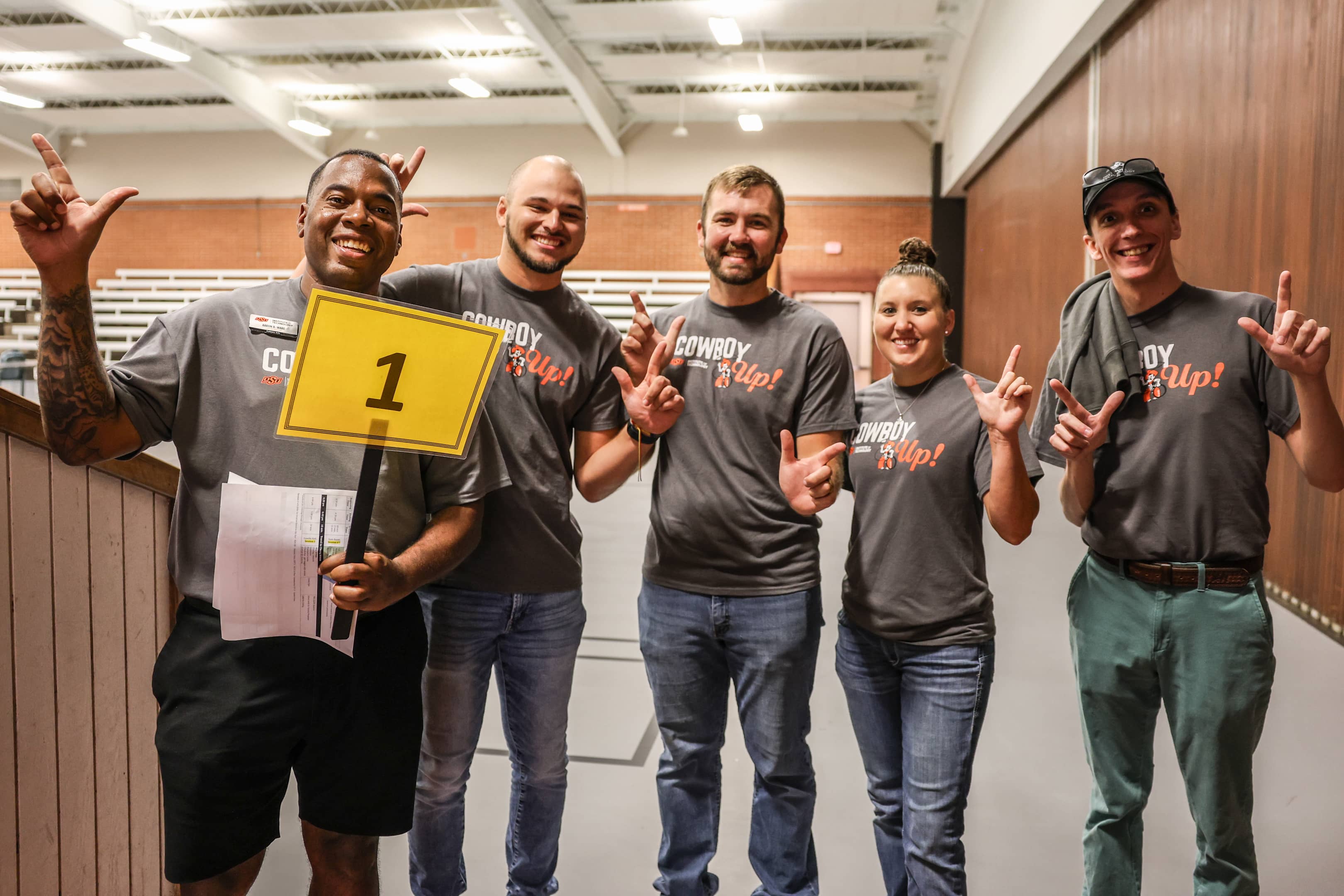 Five OSUIT team members smiling and holding a '1' sign, wearing 'Cowboy Up!' shirts at an indoor event.