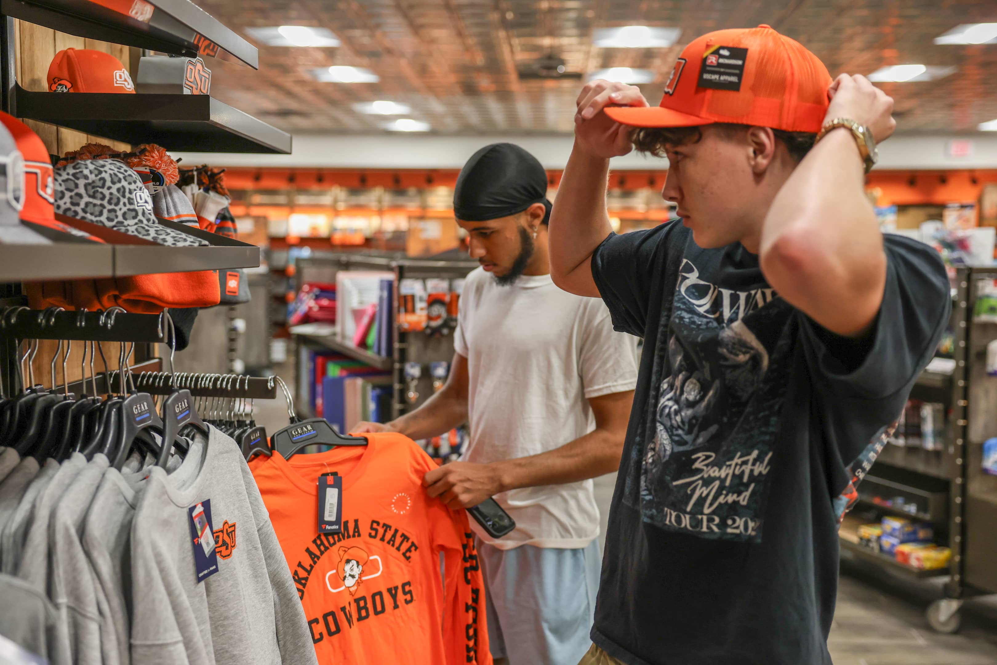 Two OSUIT students shopping in the campus bookstore, one trying on an orange cap and the other holding an Oklahoma State University t-shirt.