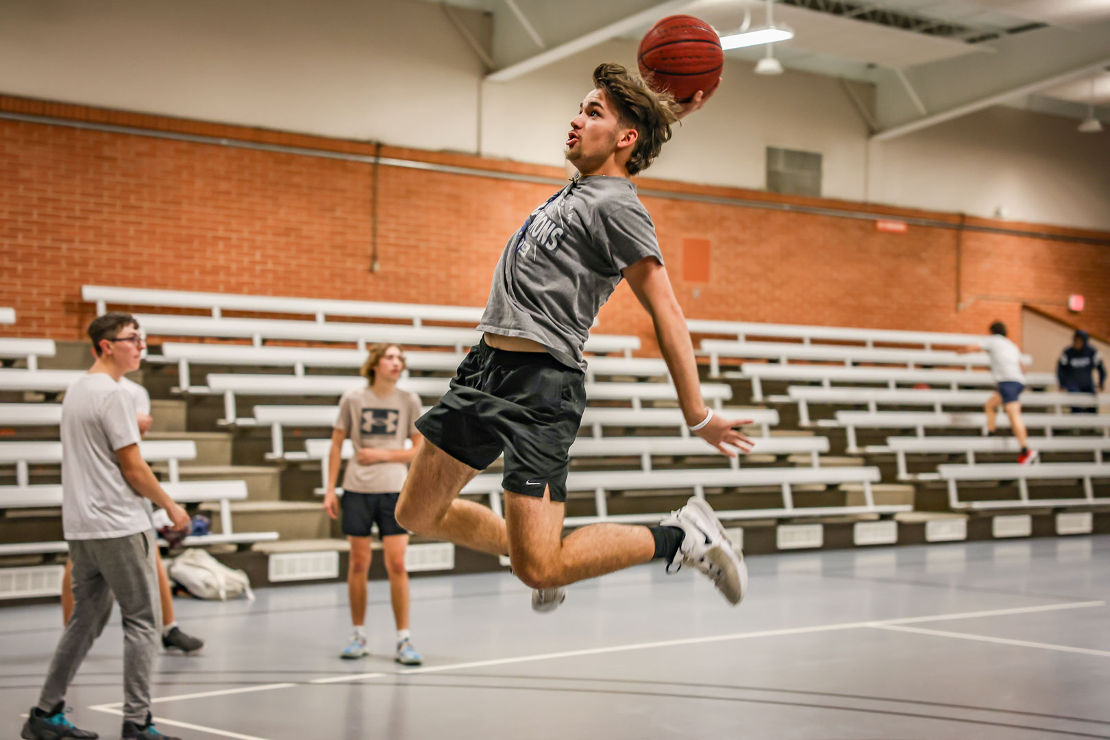 A male OSUIT student mid-air, about to dunk a basketball during a game in the gym, with other students watching in the background.