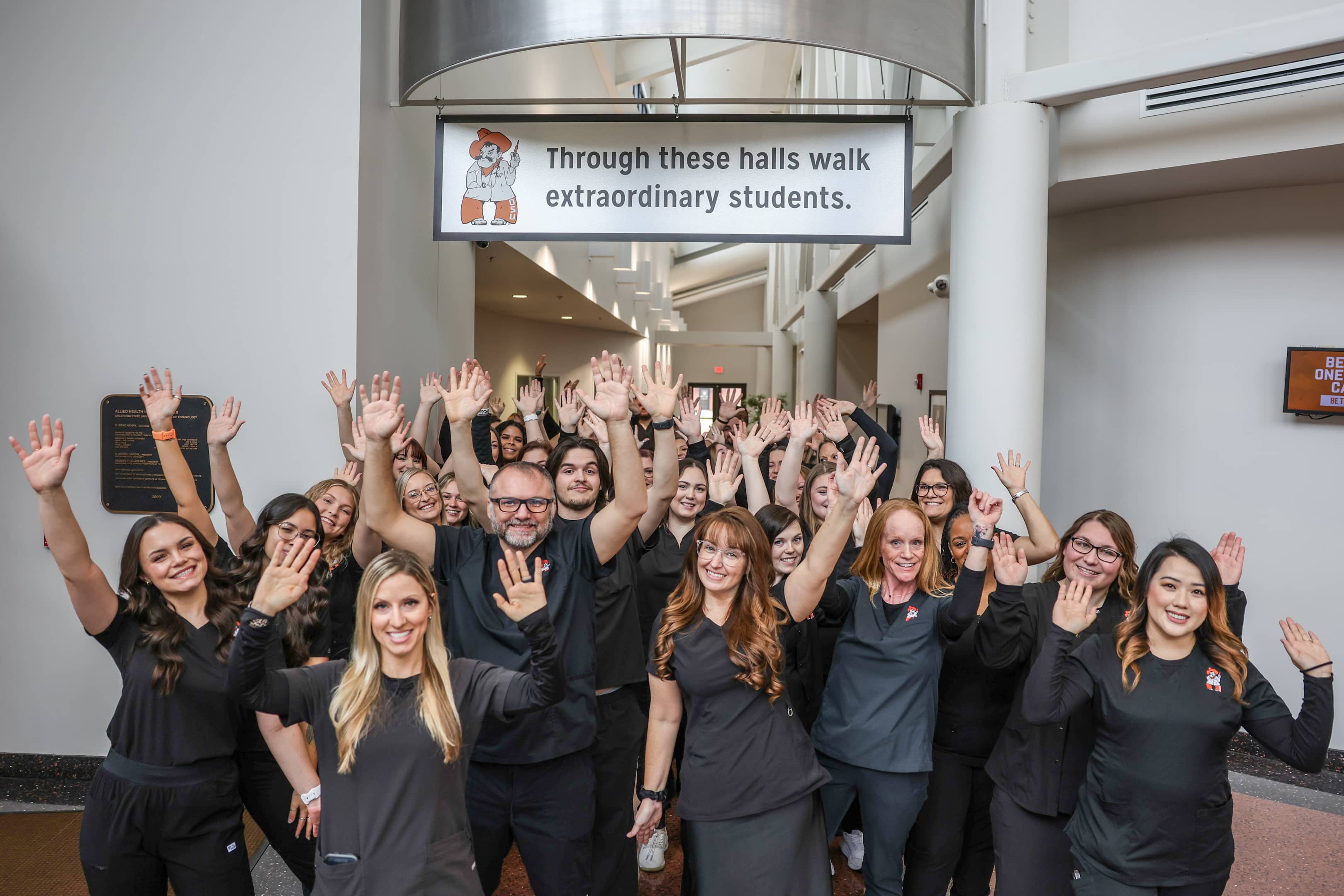 A large group of OSUIT students and staff smiling and waving under a sign that reads 'Through these halls walk extraordinary students.'