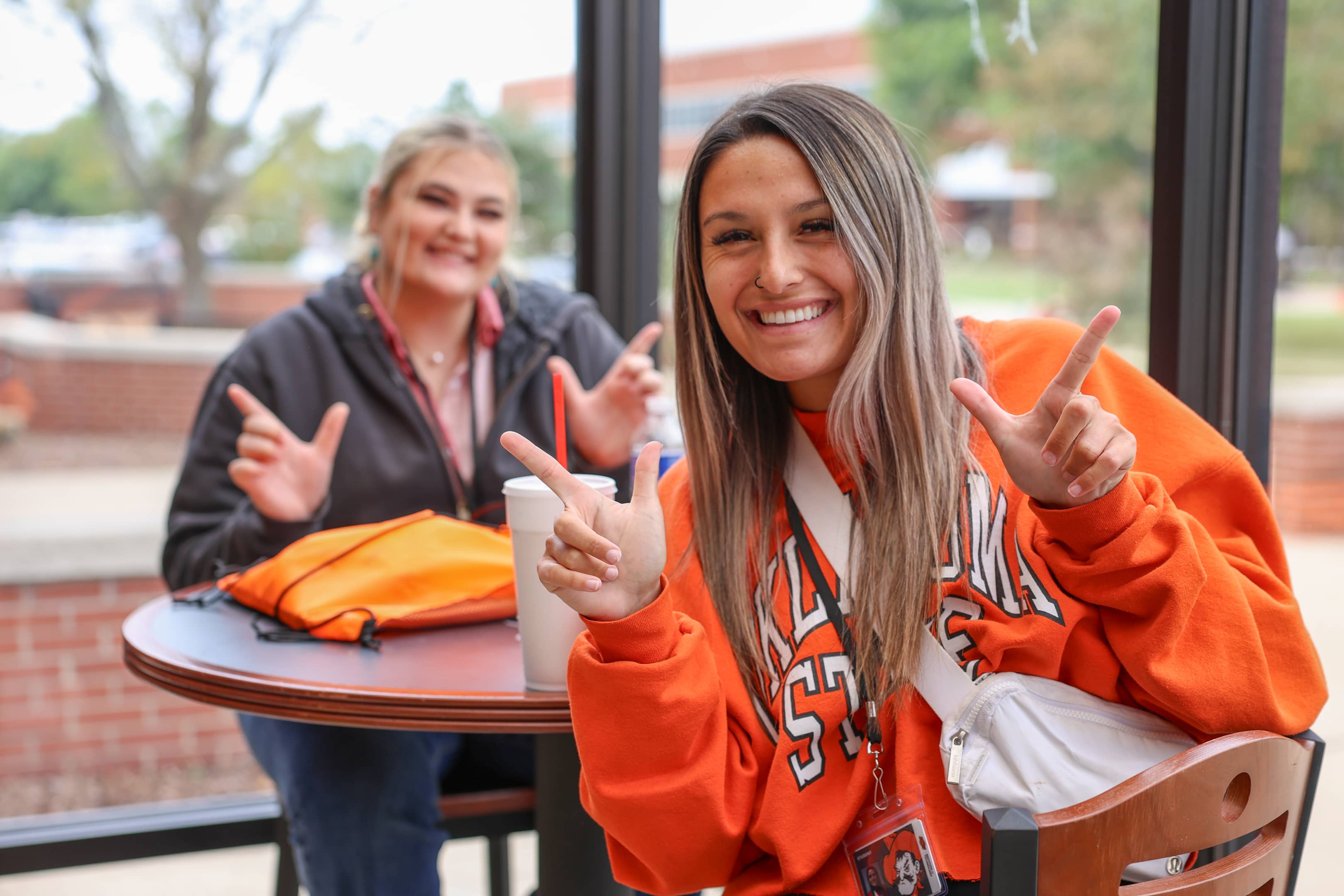 Two female OSUIT students smiling and making 'Pistols Firing' hand signs while sitting at a table, showing school spirit.