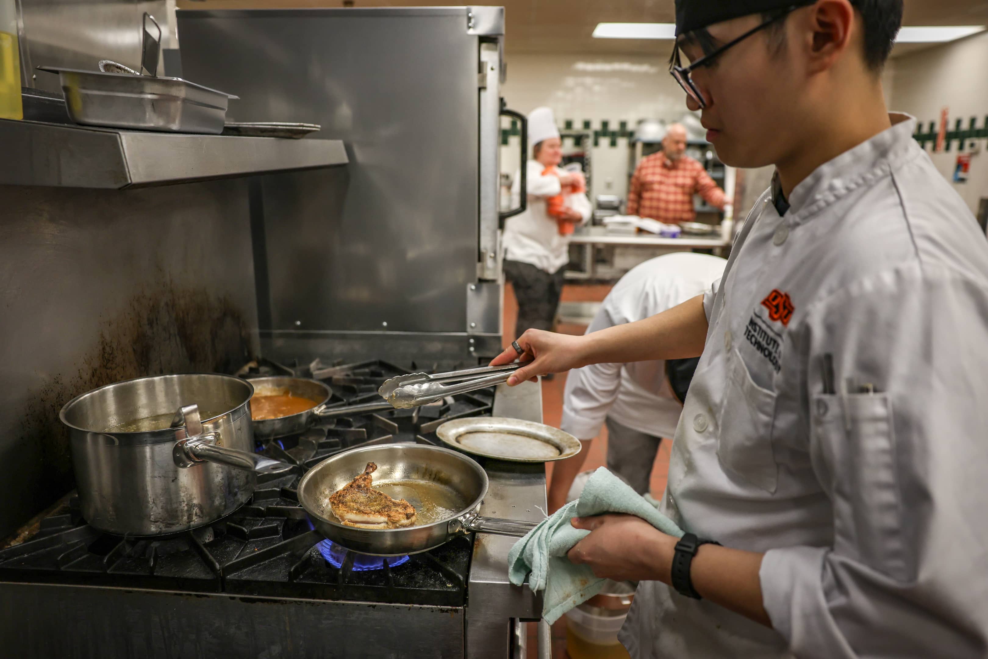 An OSUIT culinary student cooking at a stove, focusing on a dish in a frying pan, with other students and instructors in the background.
