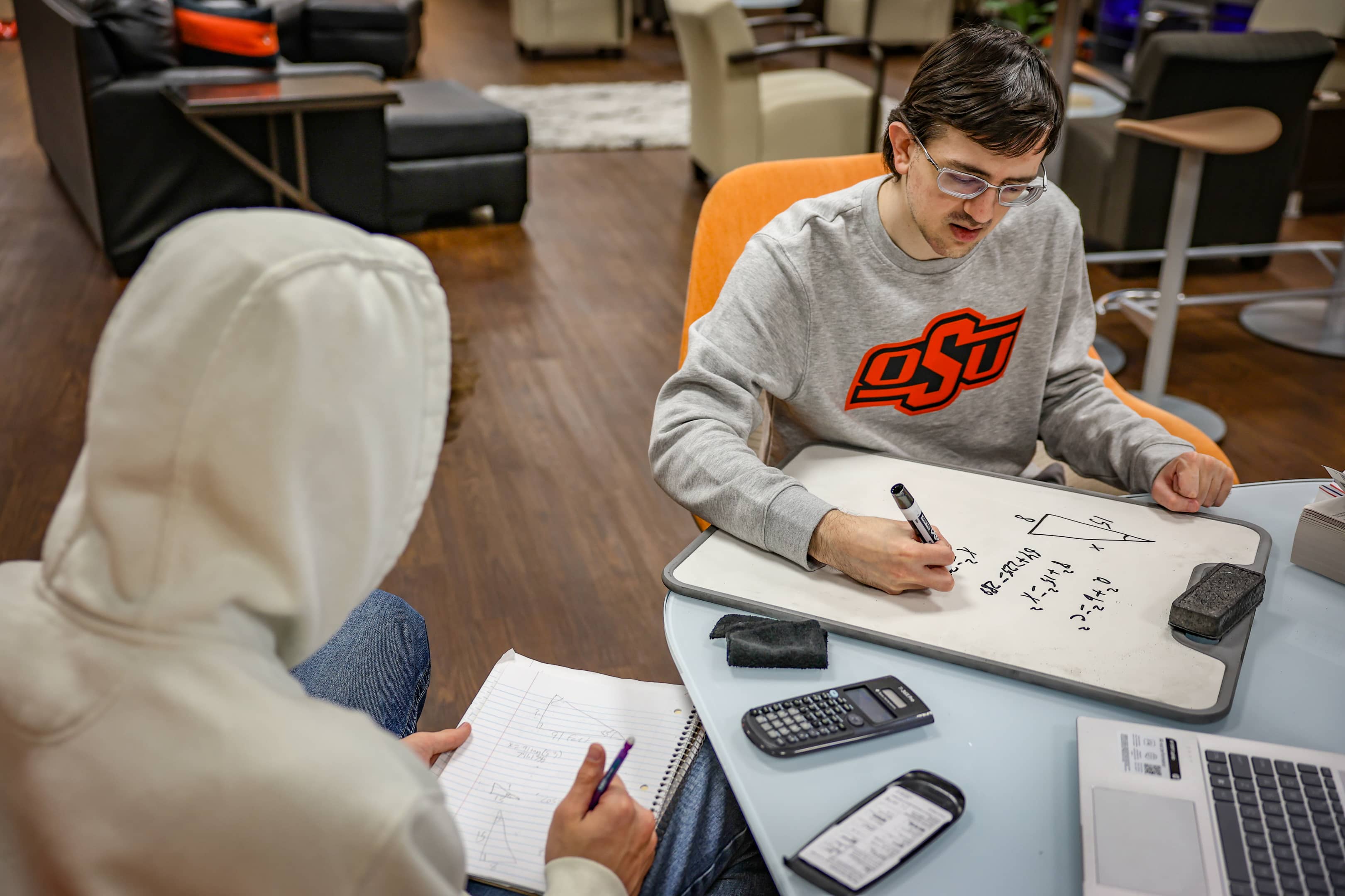 Two OSUIT students working on a math problem together in the LASSO Center, with one writing on a whiteboard and the other taking notes.